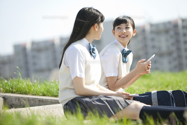 土手に座って話をする女子校生2人 の写真素材 イラスト素材 アマナイメージズ