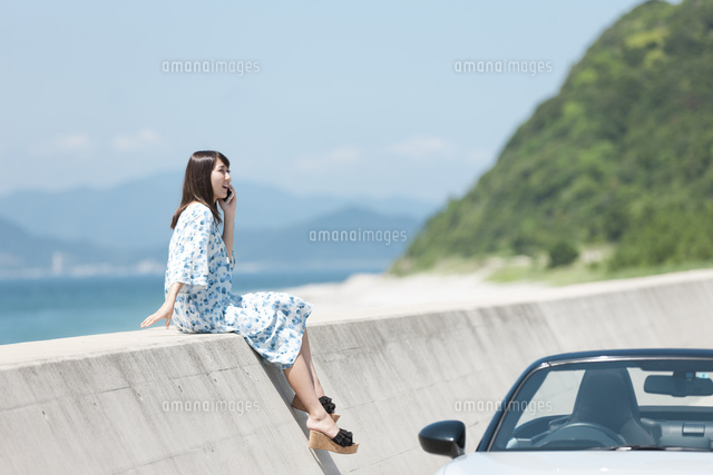 防波堤に座って電話する女性と車 の写真素材 イラスト素材 アマナイメージズ