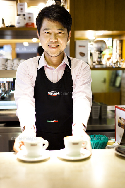 カフェのカウンター越しにコーヒーを両手にサービスをする笑顔の店員 の写真素材 イラスト素材 アマナイメージズ