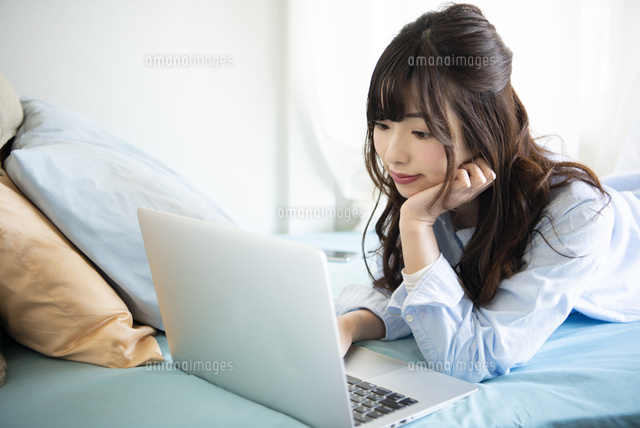 ベッドに寝転がってノートパソコンを見ている女性 の写真素材 イラスト素材 アマナイメージズ