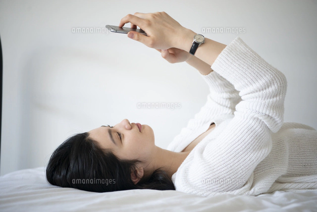 ベッドに寝転んでスマホを触っている女性[10568007832]の写真