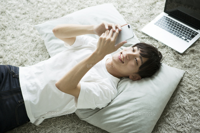 部屋で寝転がりながらスマホを操作する代男性 の写真素材 イラスト素材 アマナイメージズ