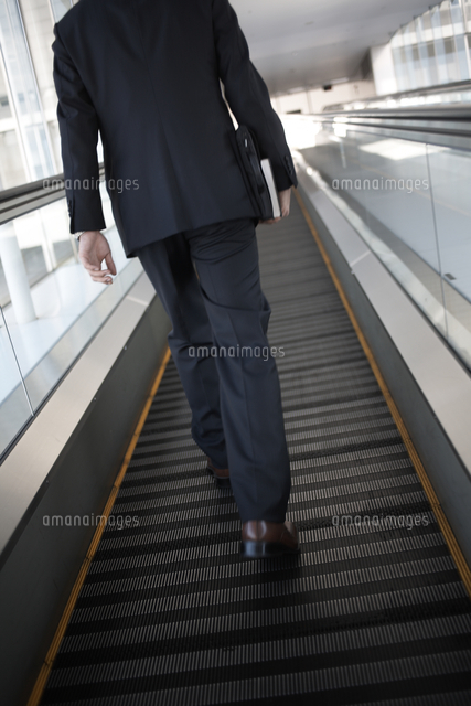 動く歩道を歩くビジネスマンの後姿 の写真素材 イラスト素材 アマナイメージズ