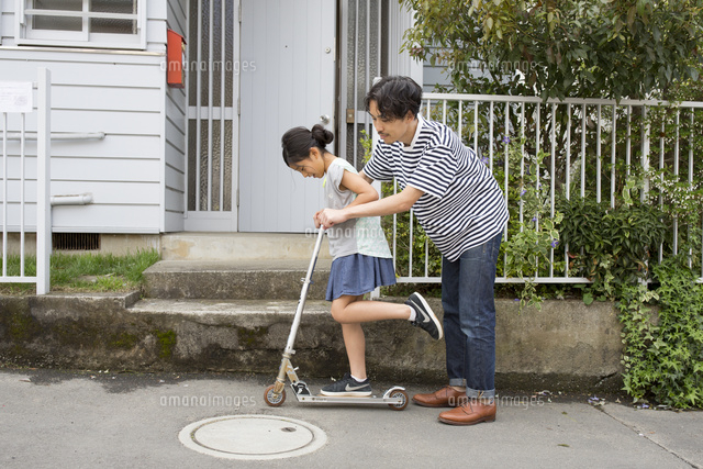 家の前でキックボードの乗り方を娘に教える父親 の写真素材 イラスト素材 アマナイメージズ
