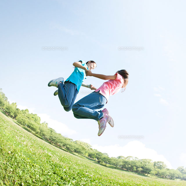 草原で手を繋いでジャンプする二人の女の子 の写真素材 イラスト素材 アマナイメージズ