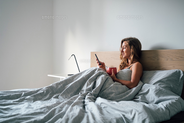 Young Woman Sitting Up In Bed Looking At Smartphone の写真素材 イラスト 素材 アマナイメージズ