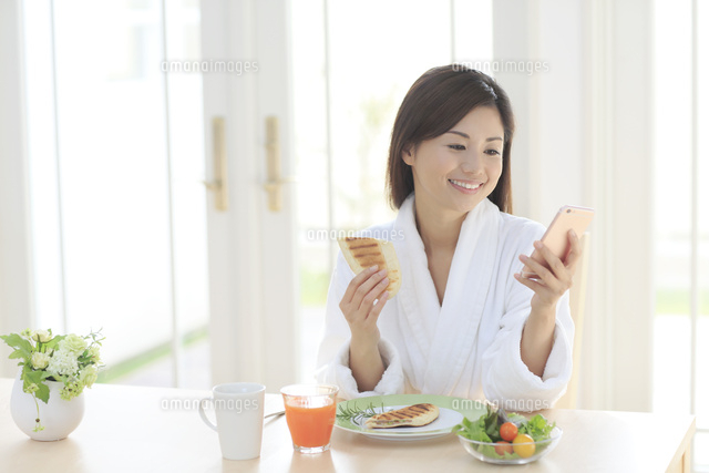 朝食を食べながらスマートフォンを持つバスローブ姿の女性 の写真素材 イラスト素材 アマナイメージズ