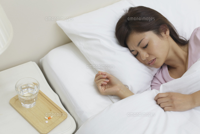 体調不良で寝る女性とテーブルに置かれた薬 の写真素材 イラスト素材 アマナイメージズ