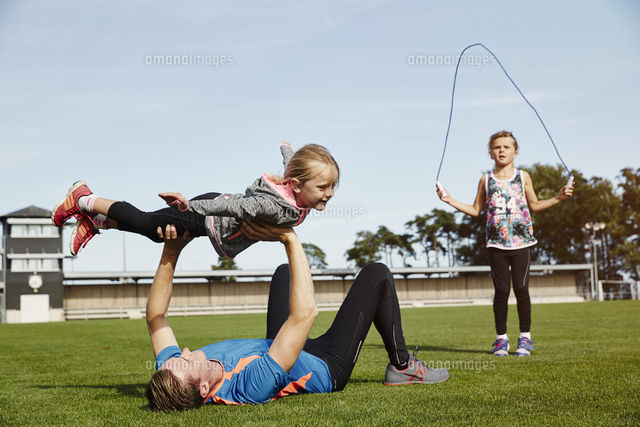 Man Lifting Daughter While Lying On Field With Girl Exercising With Jump Rope Against Clear Sky の写真素材 イラスト素材 アマナイメージズ