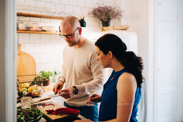 Woman With Wineglass Looking At Male Friend Cutting Pepper In Kitchen の写真素材 イラスト素材 アマナイメージズ