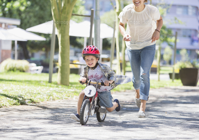 Mother chasing son riding bicycle with helmet in sunny park 