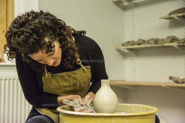 Woman with brown curly hair wearing apron shaping clay vase on pottery ...