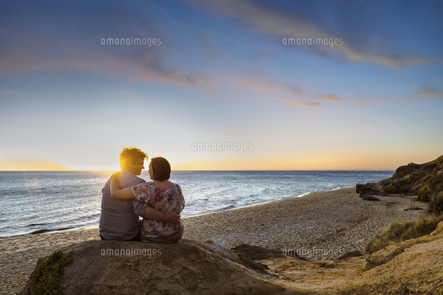 Rear view of couple sitting on rock at beach against sky during sunset ...