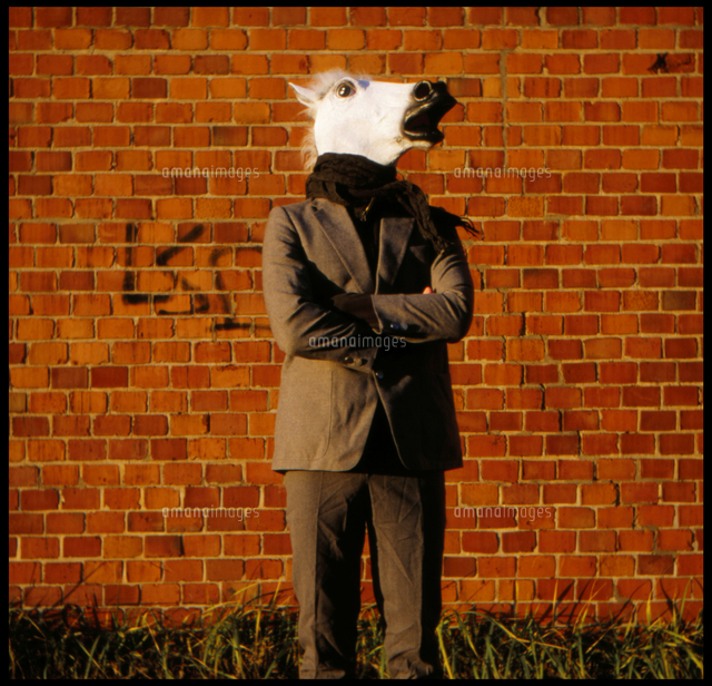 man wearing horse mask standing with arms crossed against brick wall