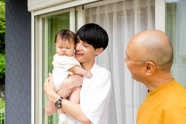 屋外の家の前で0歳児の赤ちゃんを抱っこする父親とそれを見守る祖父の3